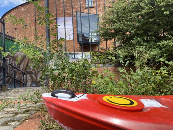 Picture of one of Roundhouse's red kayaks with its Roundhouse logo painted at the front. the kayak is on the path with the Roundhouse in the background. Picture taken on a sunny day.