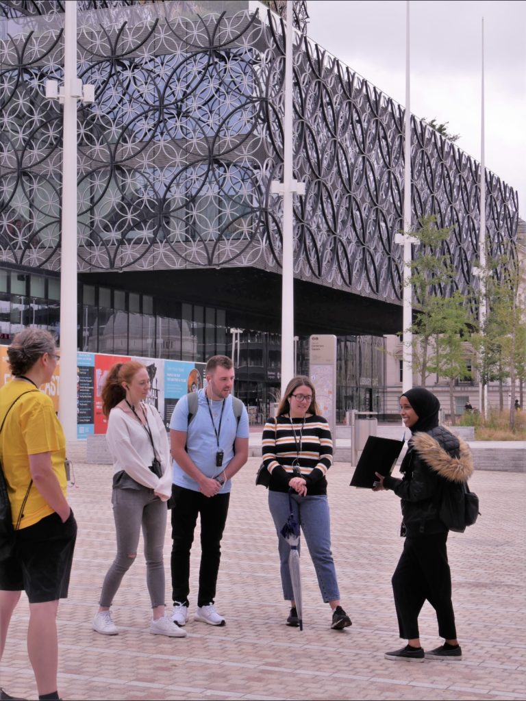 Picture of three visitors on a Roundhouse City in One Square Guided Walk, standing outside Birmingham Library looking at pictures in a folder