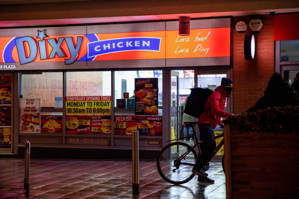 Photograph of cyclist on a rainy night going past an illuminated Dixy Chicken takeaway. The photo is called Bike by Ahsen Sayeed and is part of the Night Photography Series