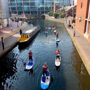 Roundhouse paddleboarders on the Birmingham canal near Symphony Hall Birmingham. The paddleboarders are looking up at the camera and smiling.