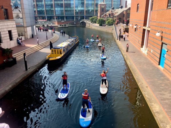 Roundhouse paddleboarders on the Birmingham canal near Symphony Hall Birmingham. The paddleboarders are looking up at the camera and smiling.