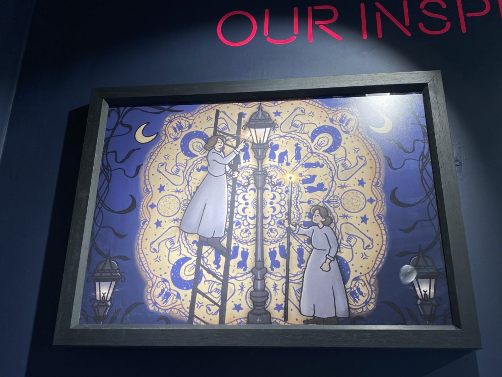 Picture of the painting Light the Way with Strength by Beth Bennett. It depicts two women in Victorian long dresses . One woman is on a ladder lighting the gas lamppost whilst the other stands by the lamp post with a fire light taller than her . The Painting uses yellow and blue and black on a blue dark background with the moon shining on both sides of the painting
