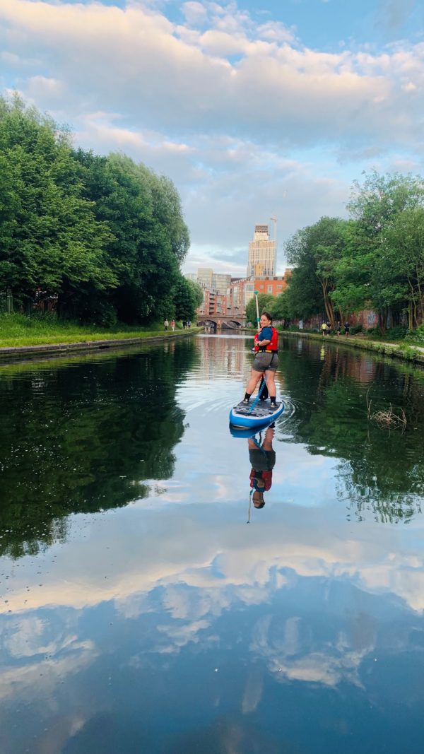 Paddleboarder on a sunny day on the Birmingham canal heading towards the city . The water reflects the clear blue sky. On both side of the canal lining the wide canal there are green trees. In the distance ahead of the paddleboarder thee are Birmingham city centre buildings