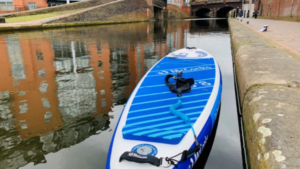 Picture of a blue and white inflated paddleboard on the canal outside the Roundhouse on a sunny day. The buildings reflect on the water.