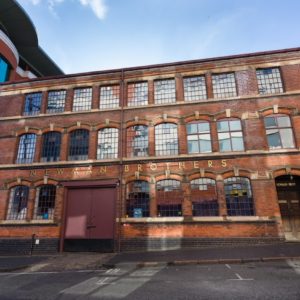 Picture of the Coffin Works factory building on a sunny day. A two floor rectangular brick building with twenty four large windows and two entrances . The Newman Brothers sign in gold in capital letters displayed across the length of the building