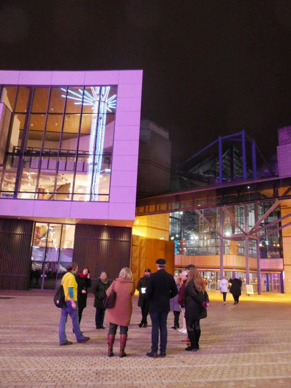 A group of people and a Roundhouse guide outside Symphony Hall and Birmingham Convention Centre taking part in the Birmingham walking tour at night.