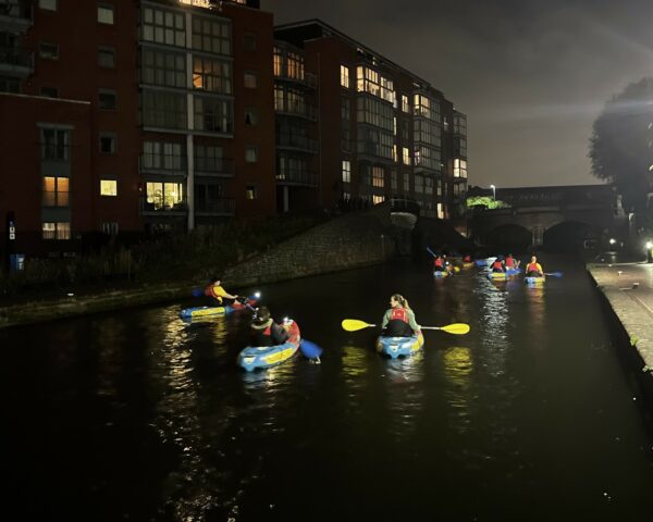 A landscape image showing 9 people on blue and red kayaks on a canal at nighttime. The kayakers are holding ores, some are blue and others bright yellow. In the background is a large apartment building light coming out of some windows.