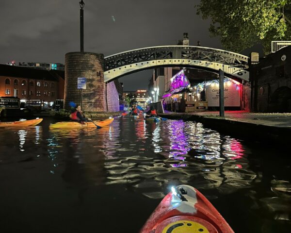 A landscape image showing a group of people kayaking on canal water. In the bottom of the image, the tip of a red and white kayak is visible with a yellow circle on it. There is a large black and white arched bridge over the canal in the distance. There is a bar on the right of the image on a path with purple and red lights, all are reflected in the canal water below.