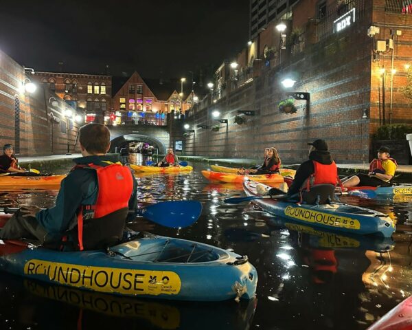 A landscape image showing a group of people sat in kayaks on canal water. Some kayaks are red, and orange and others are red and blue. Everyone is wearing a red buoyancy aid. In the distance is an arched bridge. There are lights around the canal, and they are reflected in the canal water.