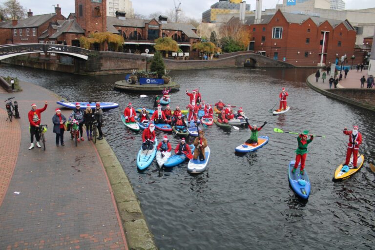 A group of people dressed as Santas and elves on stand up paddleboards, with bikes and in kayaks at Old Turn Junction on the canal
