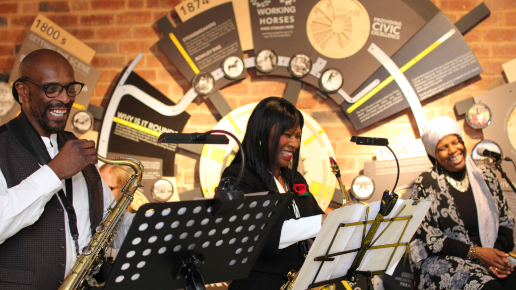 Three black people in the Roundhouse smiling playing instruments at My Voice My Path Audio Tour Launch.