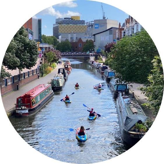 Round picture of five kayakers on the canal on a clear day. They are kayaking between two rows of moored narrowboats