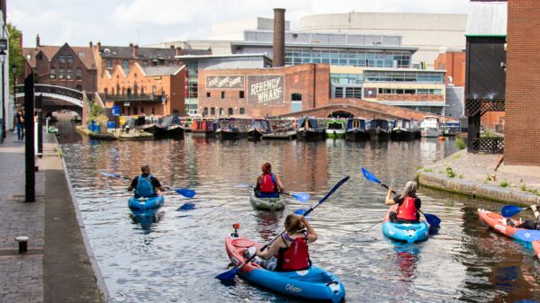 The back of a group of Roundhouse kayakers in the canal. There are canal boats moored across the canal and buildings in the background