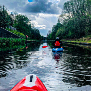 View of the canal from a kayak. There is another kayaker ahead and a large amount of trees on either side of the cut.