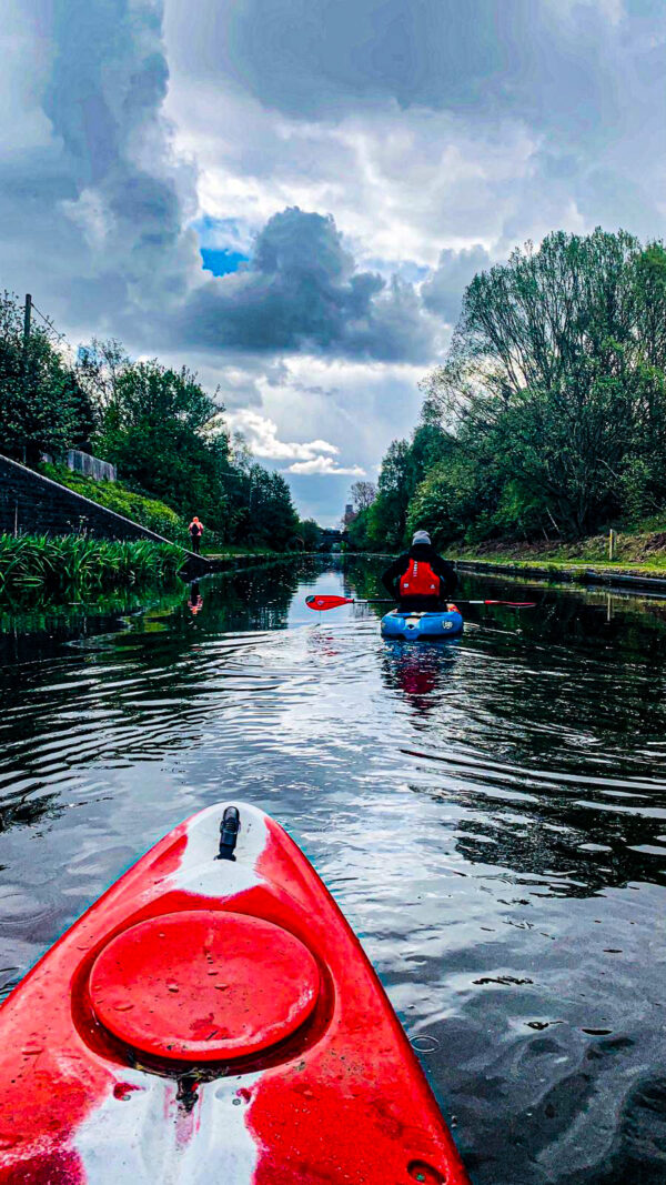 View of the canal from a kayak. There is another kayaker ahead and a large amount of trees on either side of the cut.