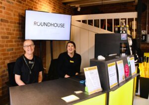 Two people smiling, wearing black behind a yellow welcome desk. There is a screen in the background showing the Roundhouse logo on a white screen