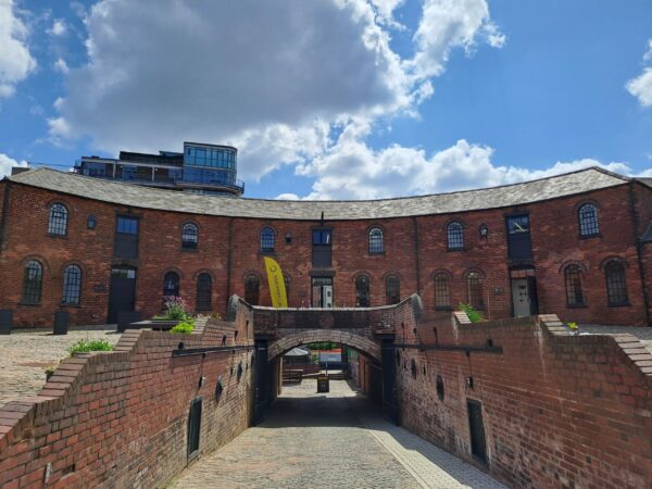 A landscape image showing a curved red brick building. In front of the image, there is a path that leads down through a tunnel. It is a blue sky day.