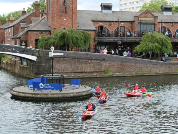 A landscape image showing people on a kayak on the canal. They are paddling towards the camera. In the background is a path and an arched metal bridge.