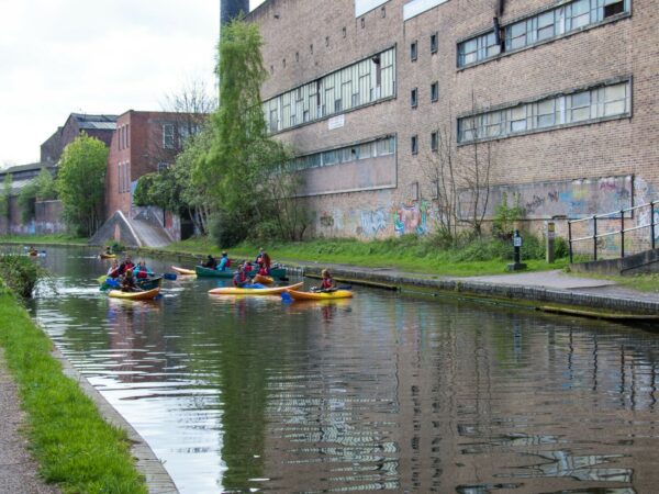 A landscape image showing a group of kayakers on a canal in Birmingham. There are lots of greenery either side of the canal. In the background is a large bricked building.