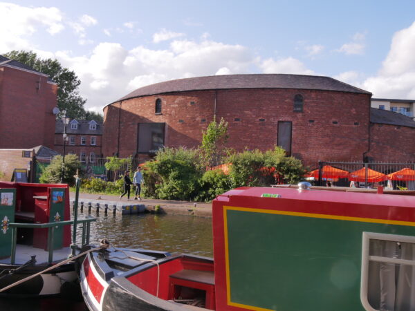 A landscape image showing the front of a red and green canal boat. In the background is an a curved red bricked building.