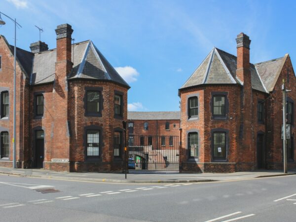 A landscape image showing a curved red bricked building on a blue sky day. In the front of the image there are two large buildings with windows.
