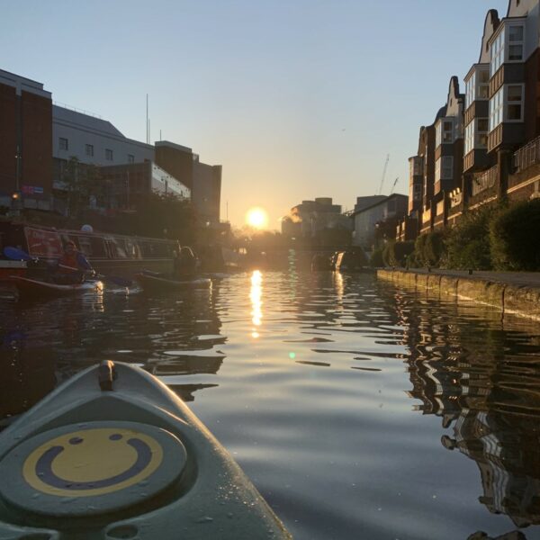 A square image showing the sun breaking and being reflecting on canal water below. To the left of the image is the front of a kayak with a smiley face logo on.
