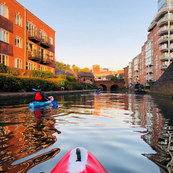 A square image showing the front of a red and white striped kayak on canal water at dawn. In front of them and to the left is another person kayaking.