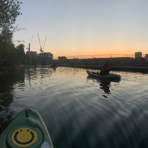 A square image showing the front of a green kayak with a circular smiley face logo on. To the right of the image is another kayaker. Behind them there is the sunrise with a gradient of orange into yellow into green into blue.