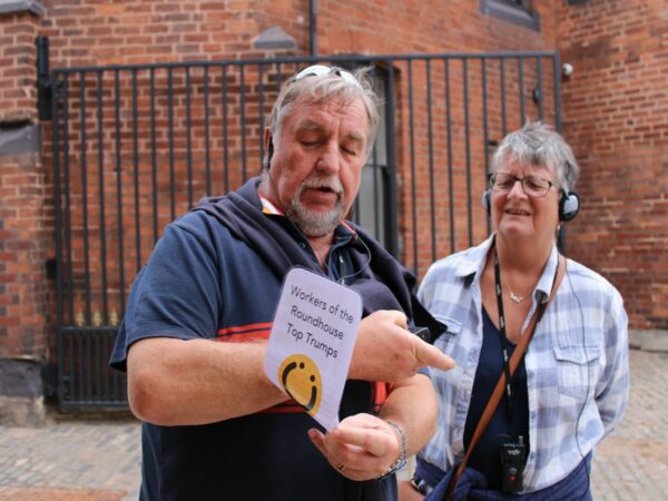 A landscape image showing two people reading information off a white card. The card has the words, 'Workers of the Roundhouse Top Trumps' written on it. Underneath the lettering is a yellow circle with a black smiling face in it. Behind them both is a black metal gate and red brick wall.