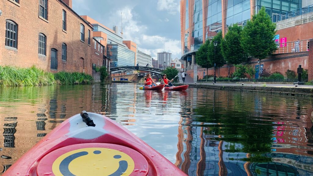 Photo taken from perspective of a kayaker of two other people kayaking on the canals. Buildings are in the background, as well as a traditional black and white footbridge