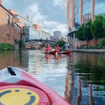 Photo taken from perspective of a kayaker of two other people kayaking on the canals. Buildings are in the background, as well as a traditional black and white footbridge