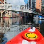 A kayak drifts towards a bridge over the canal with large apartments all around, the Roundhouse yellow smiley face logo on front of the kayak can be seen in the middle of the photo