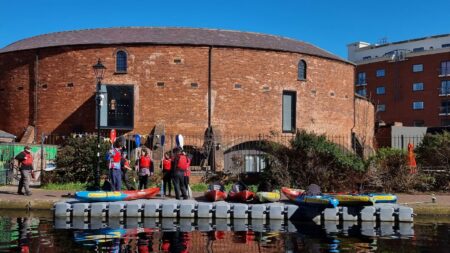 A landscape image showing a red bricked curved building on a bright blue skied day. In the foreground is a canal and on the canal is a pontoon. Stood next to the pontoon on a path are a group of people and some red and blue kayaks.