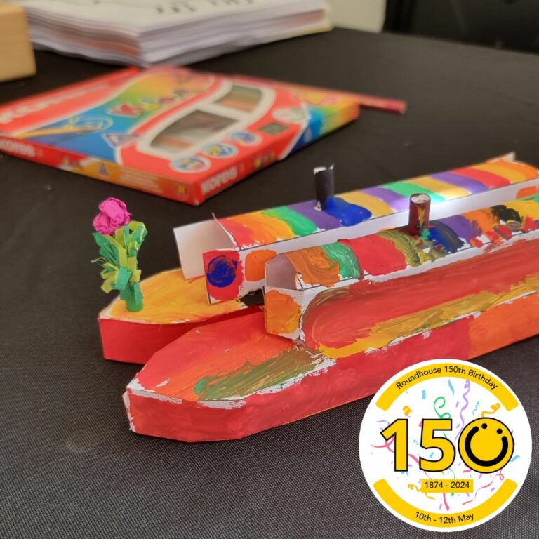 A square image showing a table with two paper canal boat models. They have been coloured in and are red, orange, yellow and blue. In the bottom right of the image is a graphic logo with the number 150, the 0 of the logo is a yellow smiley face logo. The numbers 1 and 5 both have a black outline.
