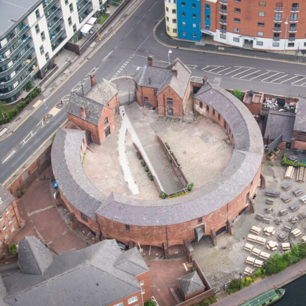 A square image showing a birds eye view of a curved building. The building is red bricked and the roof if grey. In the centre is a cobbled courtyard, with a tunnel and a lighter grey paved path curving around on the left of the image.