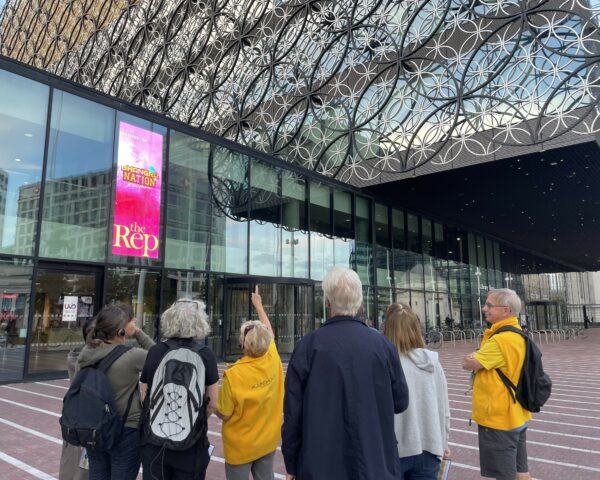 A landscape image showing a group of people looking up at a glass building. The building is silver and yellow with silver metal overlay. The group of 7 people are stood looking up and one person, wearing a yellow shirt is pointing up at the building.