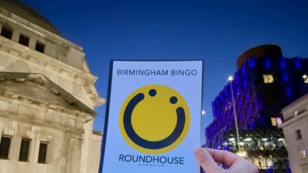 A landscape image showing a hand holding a piece of paper with a yellow circle smiley face on with the words ‘Birmingham Bingo’ above it and ‘Roundhouse Birmingham’ underneath it. To the left is a white building and to the right is a building let with blue lights. The background is yellow. In the bottom right of the image is a yellow smiley face logo and a red square with the letter h in.