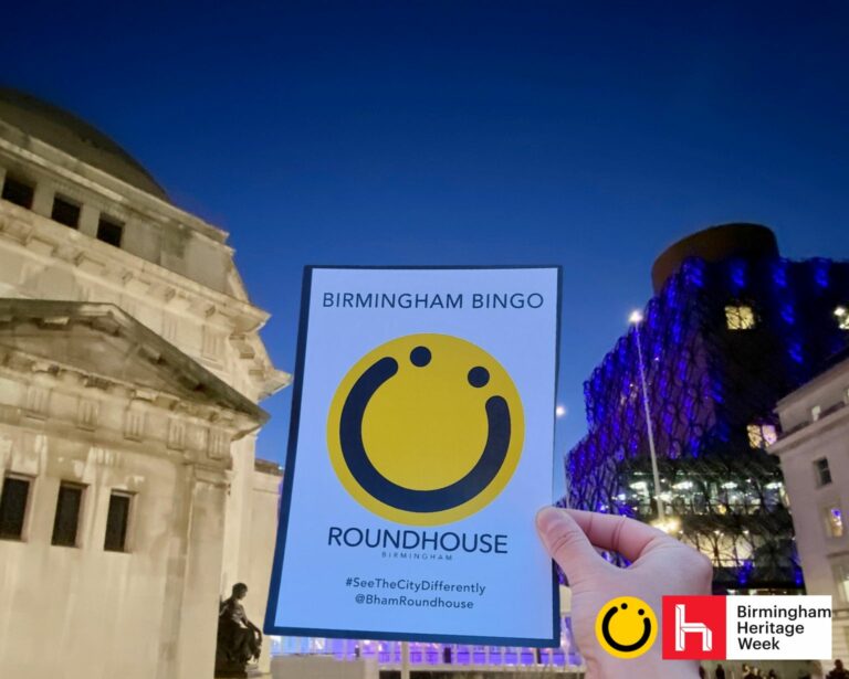 A landscape image showing a hand holding a piece of paper with a yellow circle smiley face on with the words ‘Birmingham Bingo’ above it and ‘Roundhouse Birmingham’ underneath it. To the left is a white building and to the right is a building let with blue lights. The background is yellow. In the bottom right of the image is a yellow smiley face logo and a red square with the letter h in.