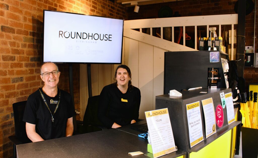 Two members of the Visitor Centre team, one male and one female, sitting at the black and yellow reception desk looking into the camera. Large tv behind them shows the Roundhouse logo on a white background.