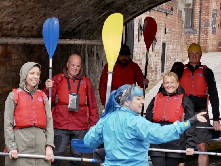 A landscape image showing someone in a blue shirt delivering a briefing to a group of kayakers. In front of them is a group of 5 people in red life jackets.