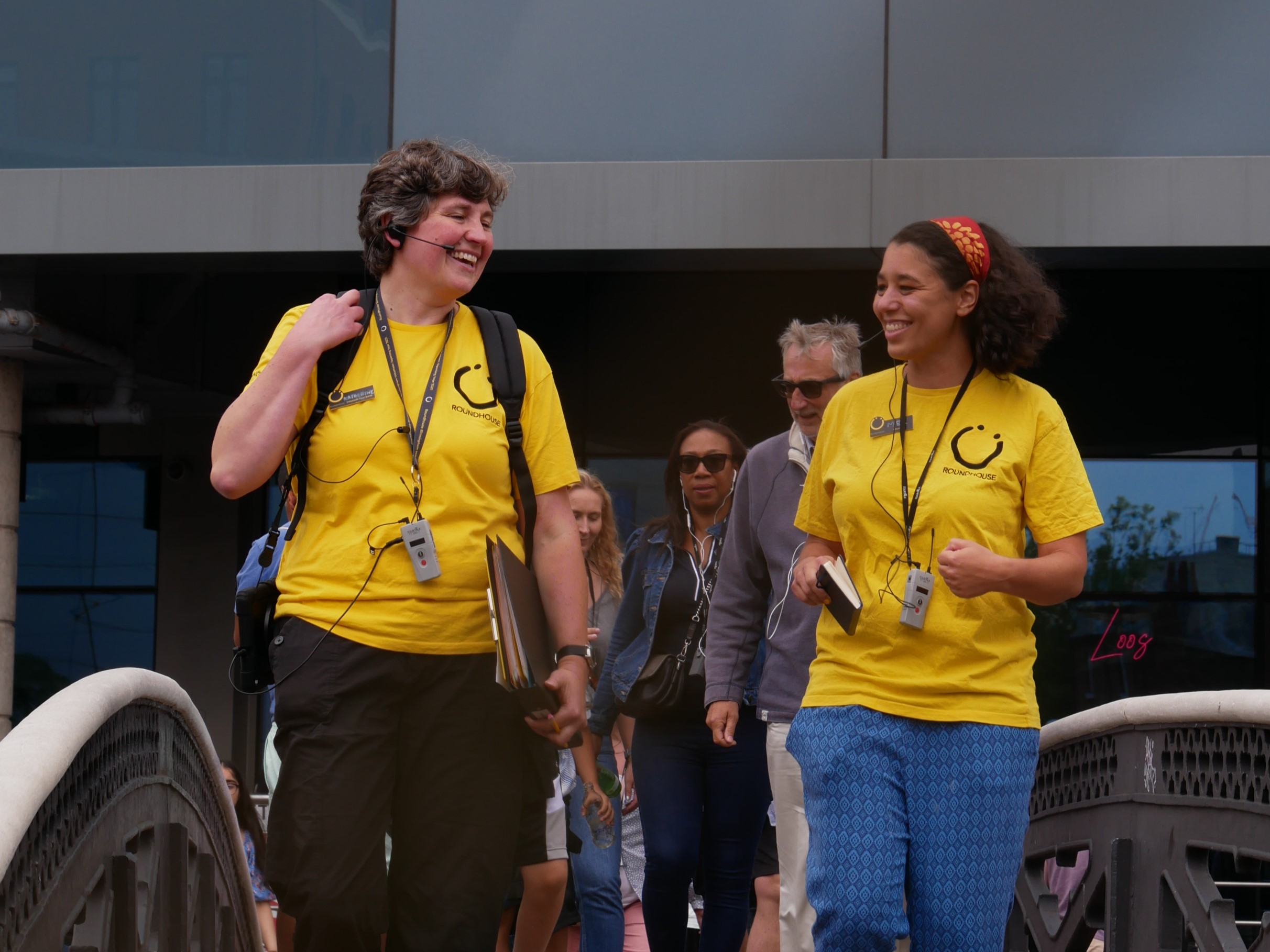 A landscape image showing two people in yellow t-shirts leading a group of people over a bridge. They are laughing and smiling at each other.