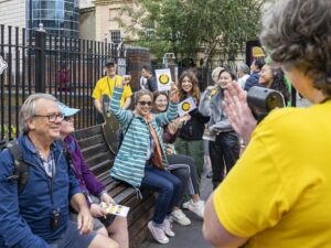 A landscape image. In the right of the image is a persons shoulder, they are wearing a yellow t-shirt. The main body of the image is a group of people holding cards with a black smiling face on a yellow background. They are cheering and have their arms up in the air, smiling.