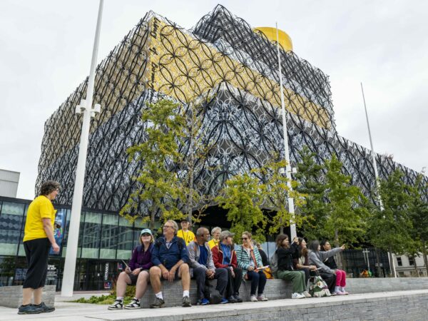 A landscape image showing a group of people sat on a concrete bench with a large silver, grey and yellow multi-layered building behind them.