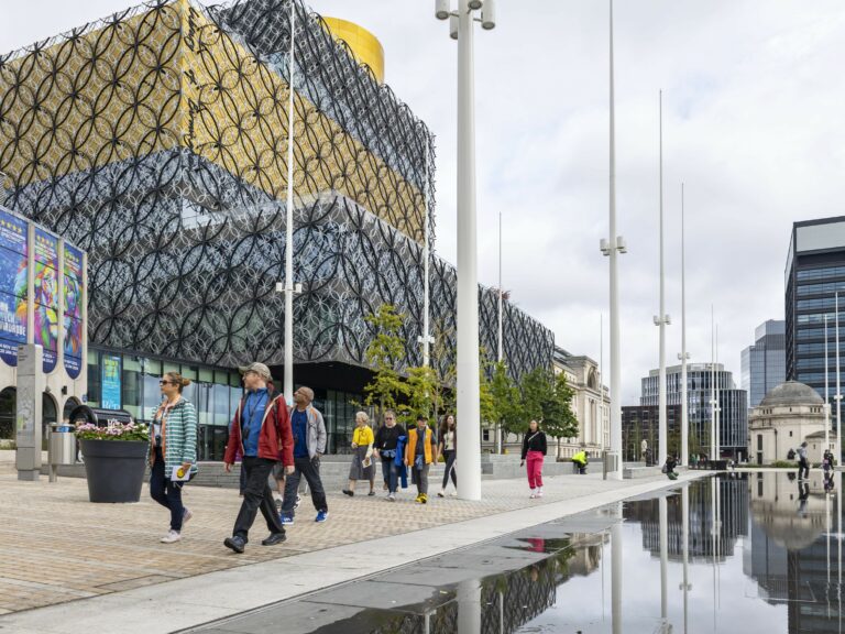 A landscape image showing a group of people walking with a large silver, grey and yellow multi-layered building behind them.