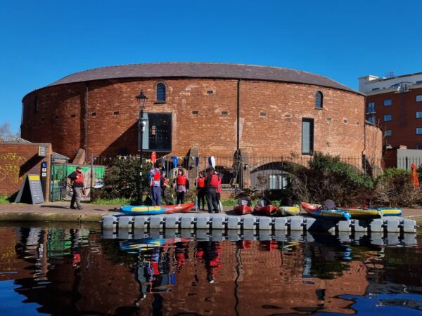 A landscape image showing a pontoon on a canal with multiple red and blue kayaks. Behind them is a curved red bricked building and a very blue sky.