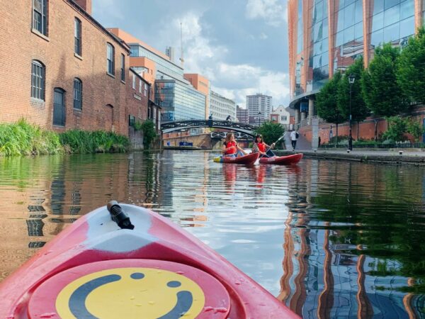 A landscape showing the front of a red kayak. In the distance are two people sat in kayaks on the canal. In the background is an arched bridged. On the front of the kayak is a yellow circular logo with a black smiling face.