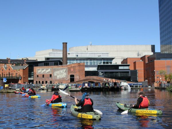 A landscape image showing a group of kayakers on a body of water. In front of them are some working canal boats and red bricked buildings.