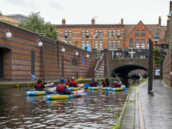 A landscape image showing a group of kayakers on a canal. The photographer stood to the right on the towpath is behind them and they are about to go through a arched tunnel. The kayaks are red and blue.
