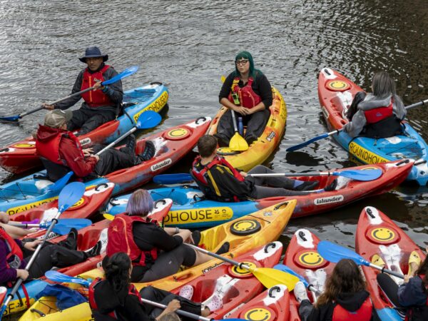 A landscape image showing a large group of people sat in kayaks on the canal water. All the kayaks are close to eachother. Its very colourful, lots of blues, reds, oranges and yellows with kayak oars in the air.