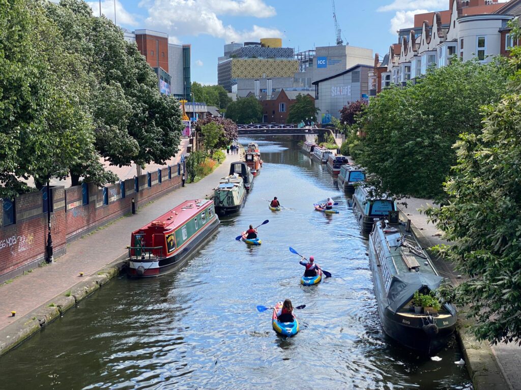 Group of kayakers on the Bustling Birmingham tour kayaking between two rows of moored narrow boat going towards Sea Life Birmingham.